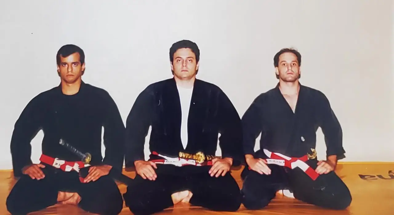 Três praticantes de quimono preto e faixa vermelha e branca, sentados lado a lado em seiza, as mãos sobre as coxas e a espada na cintura, diante de uma parede branca.