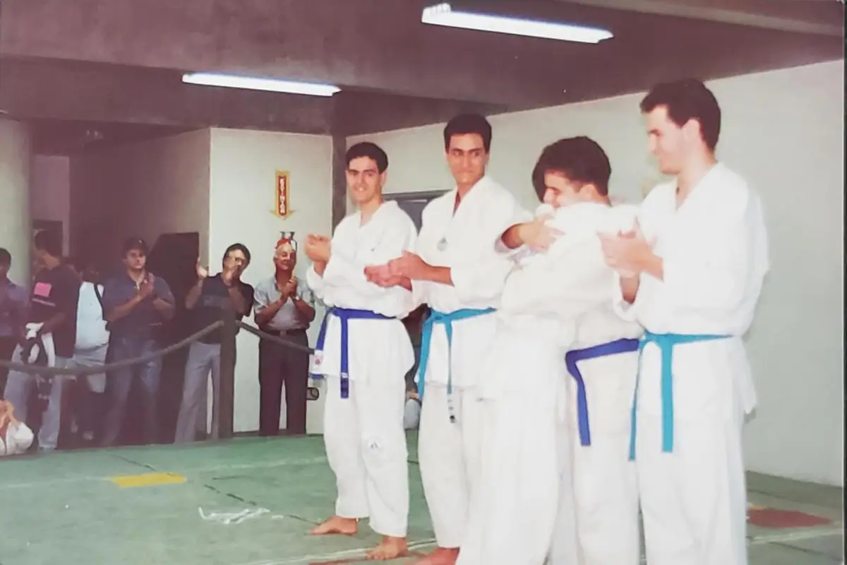 Quatro alunos de quimono branco e faixa azul se cumprimentam e aplaudem sobre o tatame, enquanto o público aplaude atrás de uma corda.